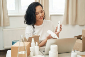 Smiling Internet shop employee holding skincare products and looking at laptop screen during video call 25th Hive