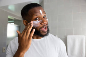Cheerful young black man lookin the camera and applying moisturizer to his face inside the bathroom at home during his morning routine at home
